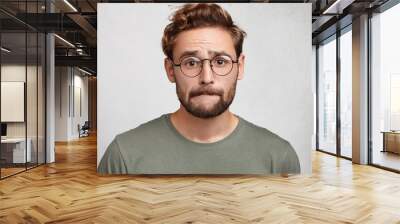 Worried bearded guy in eyewear, bites lower lip, anticipate important decision or feels nervous before doctor`s visit, being afraid of it. Uncertain attractive male nerd worries before final exam Wall mural